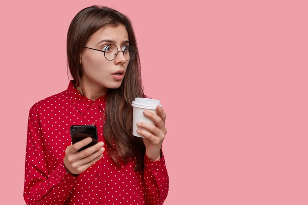 puzzled surprised dark haired beautiful woman holds take out coffee, modern cell phone, dressed in red blouse