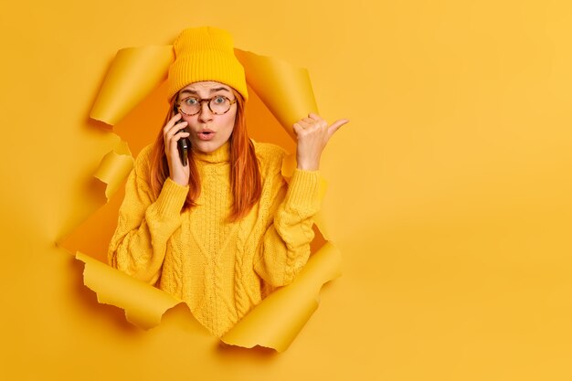 Puzzled surprised millennial redhead woman reacts on something astonishing points right with thumb dressed in casual clothes talks via smartphone breaks through paper hole