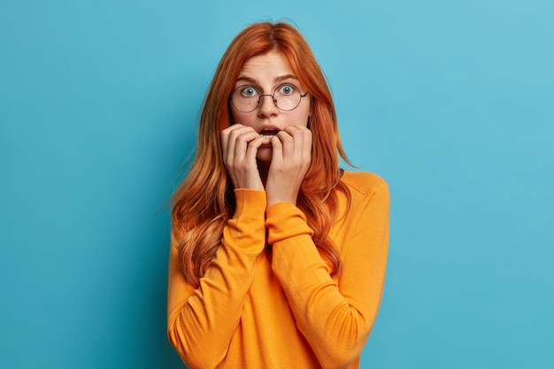 Puzzled surprised redhead Caucasian woman bites finger nails keeps mouth opened gasps from amazement dressed in casual jumper.