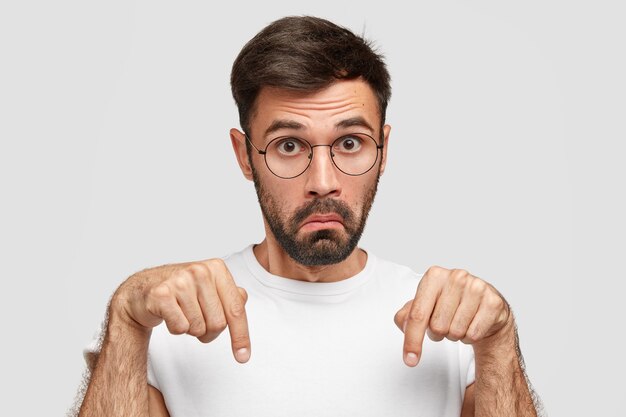 Puzzled surprised unshaven young man with dark stubble, points down with both index fingers