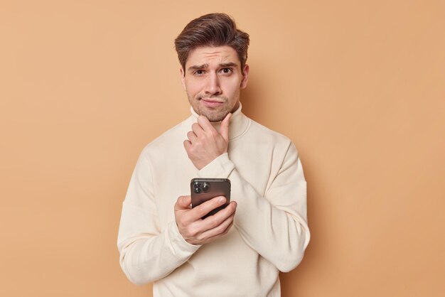 Puzzled thoughtful adult man holds chin and ponders on something holds mobile phone considers receieved notification thinks what answer to give wears turtleneck isolated on beige studio wall