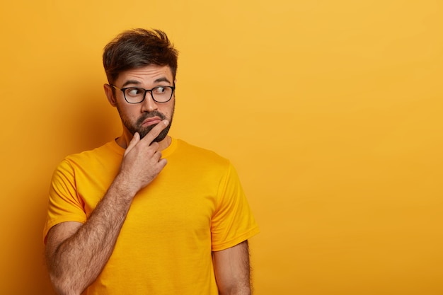 Puzzled thoughtful young man holds chin and looks away