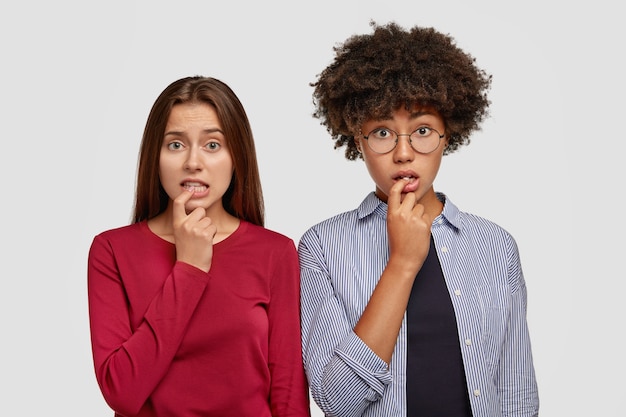 Puzzled two girls bite fingers, look with indecisive facial expression, try find solution, wear casual clothes, look with frustration, model against white wall. Diversity and reaction concept