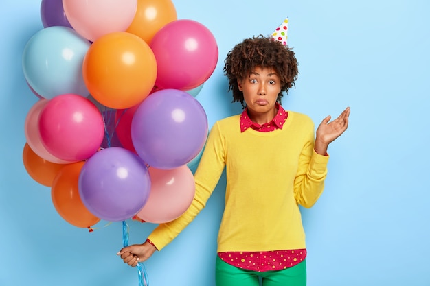 Puzzled unaware Afro woman stands with colored balloons, doesnt know where party is held