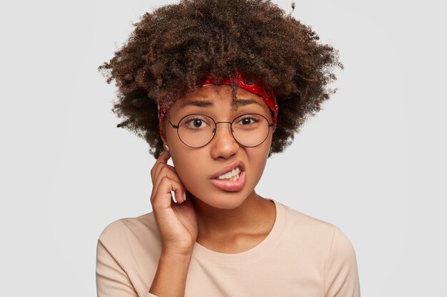 Puzzled uncertain black girl frowns face in displeasure, feels apathy, looks with uncertainty, cant find solution and way out of problem, has Afro haircut, models against white wall. Facial expression