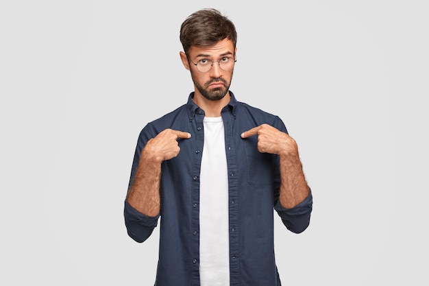 Puzzled unshaven male points at himself, looks with bewilderment, dressed in casual shirt, frowns face, wears spectacles, wonders to be chosen for giving word, isolated over white wall