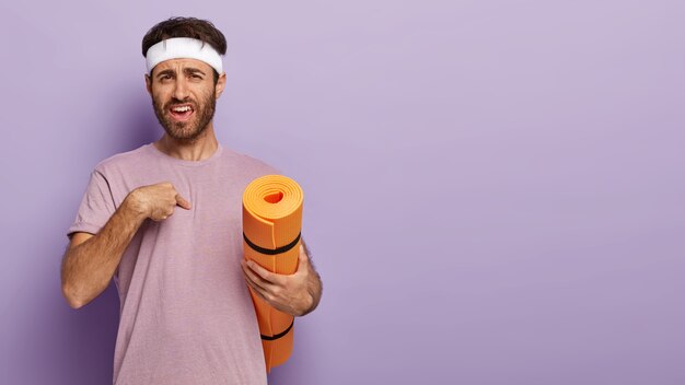 Puzzled unshaven man points at himself with indignation, wears white headband and casual purple t shirt, asks trainer whether exactly he should do exercise, carries mat