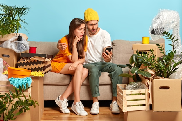 Озадаченные женщина и мужчина смотрят на смартфон, переезжают в новую квартиру, ищут мебель для своей квартиры в интернет-магазине.