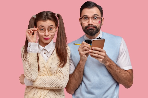 Puzzled wonk guy with lipstick on cheek, writes down information in notepad, pretty European girl keeps hand on frame of big spectacles