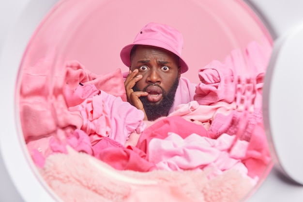 Puzzled worried Afro American man stuck in pile of laundry busy doing washing at home poses from inside of washer shocked to receive one more task from wife