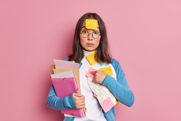 Puzzled young Asian woman points at herself asks who me looks surprisingly at camera