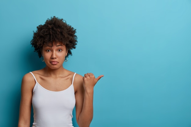 Puzzled young ethnic woman points thumb on copy space, dressed in casual vest, hesitates about product price, sees something strange, poses 