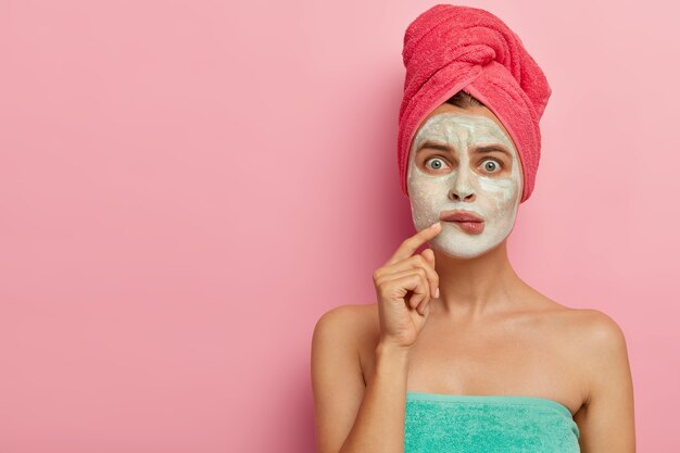 Puzzled young female bites lips, applies clay mask on face, stands topless in towel, models against pink wall, has beauty treatments, cares about herself. People, wellness and peeling concept