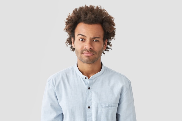 Puzzled young man with Afro hairstyle raises eyebrow in bewilderment, reacts on something, feels doubt, dressed in elegant shirt, isolated over white wall