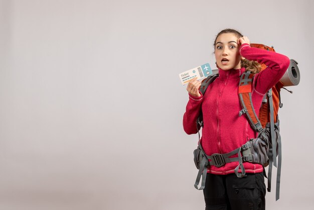 puzzled young traveller with big backpack holding up travel ticket on grey