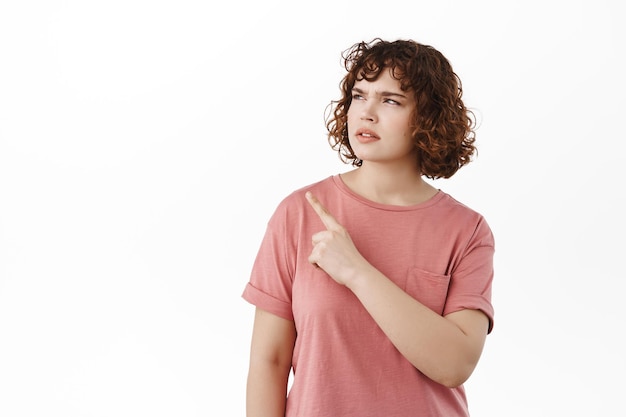 Puzzled young woman, pointing and looking aside at upper left corner promo offer, staring questioned and confused at banner advertisement, standing over white background.