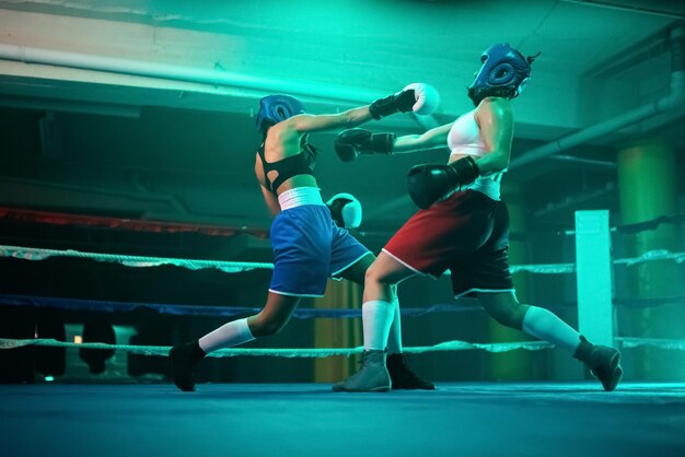 Qualified female boxers boxing in gym.  Two young girls in helmets and gloves standing in blue light on ring, aggressively attacking and punching each other. Healthy lifestyle and combat sport concept