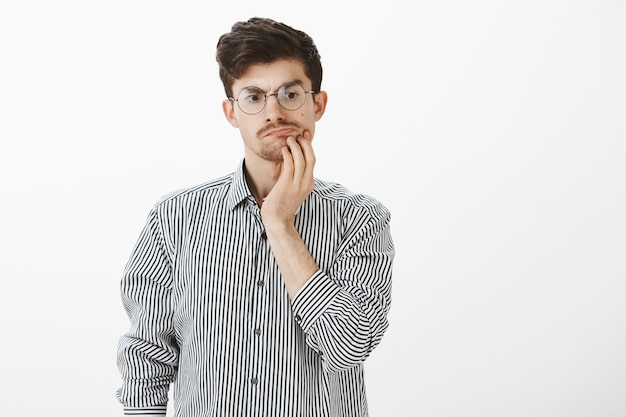 Questioned doubtful attractive bearded guy with moustache in round glasses, holding hand on chin and looking down while thinking or spacing out during conversation, being away from discussion