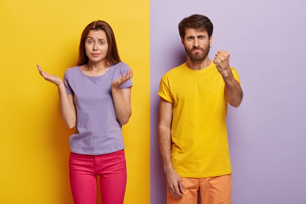 Questioned doubtful woman spreads palms sideways, has unaware facial expression, angry man shows fist at camera