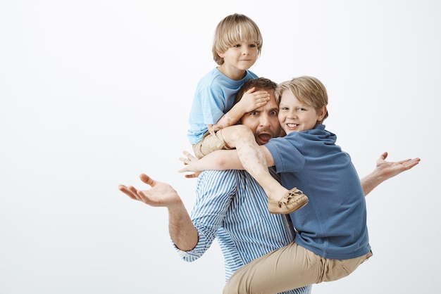 Questioned european father raising spread palms in clueless gesture sticking out tongue and looking concerned at camera while son sitting on shoulders and touching forehead and kid hanging on chest