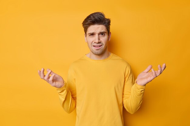 Questioned hesitant adult man spreads palms looks with bewiderment doesnt knowwhat to do has puzzled expression wears casual jumper isolated over yellow background Human perception and attitude