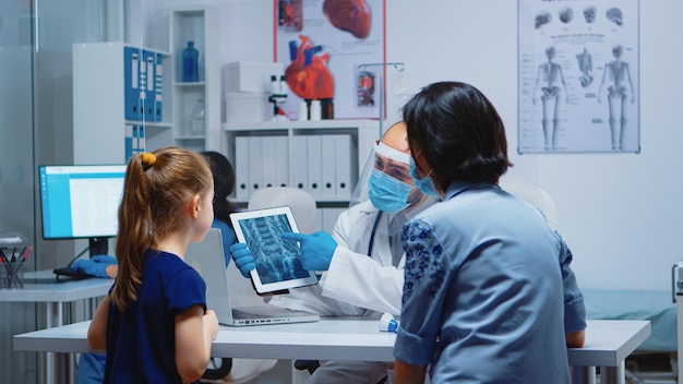 Free photo radiologist explaining x-ray using tablet in medical office and nurse working on computer. pediatrician specialist with protection mask providing health care service radiographic treatment examination