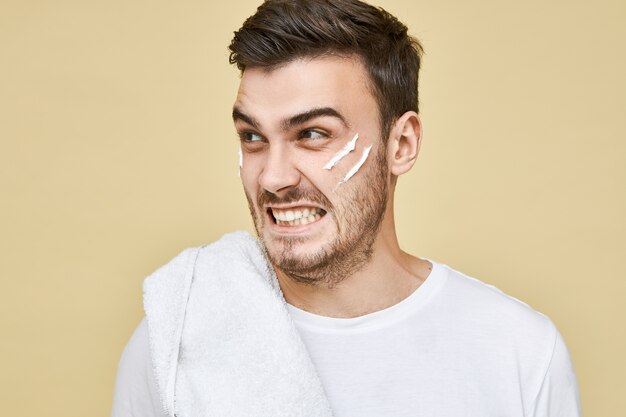 Rage and anger. Close up shot of fierce angry young man with towel in shoulder grimacing, posing  with shaving foam applied on his face, can't shave because hot water was turned off