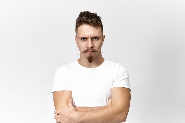 Rage, bad mood and fury concept. Portrait of handsome young man with mustache and stubble keeping arms crossed on his chest and frowning, trying to restrain his anger and negative reaction