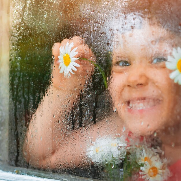 村の窓ガラスの雨滴、子供たちの手の中のカモミールの花の目が雨を見ています。