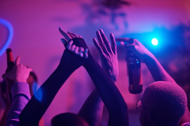 ホームパーティーで踊り 週末を楽しみながらピンクのライトに対して飲み物を飲んでいる若い興奮した異文化間の友人の腕を上げた プレミアム写真