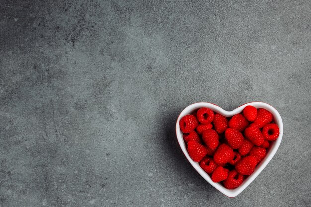 Raspberries in a heart shaped bowl on a gray textured background. top view. space for text