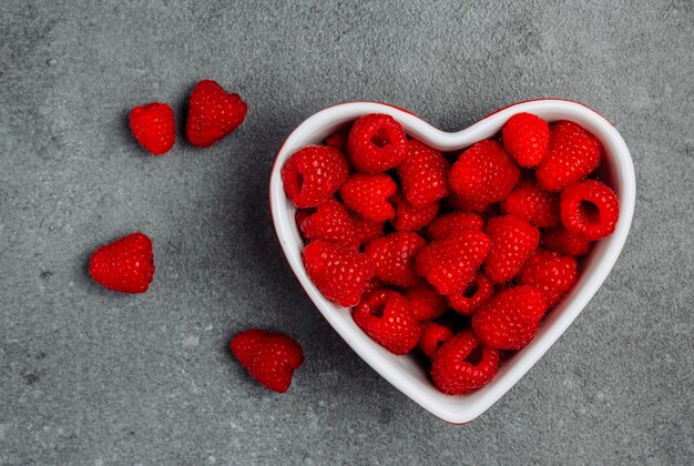 Raspberries in a heart shaped bowl on a gray textured background. top view.