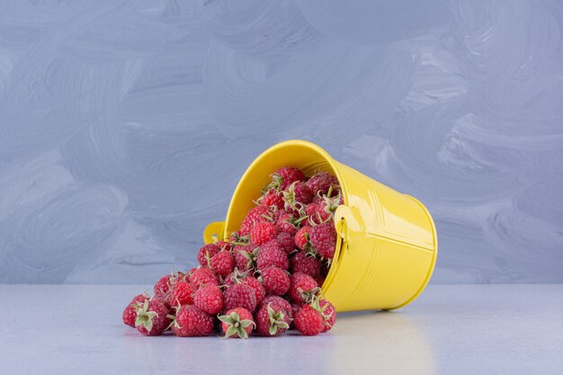 Raspberries pouring out of a yellow bucket on marble background. High quality photo