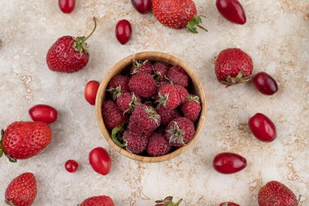 Raspberries in wooden bowl with hips and strawberries scattered on marble background. High quality photo