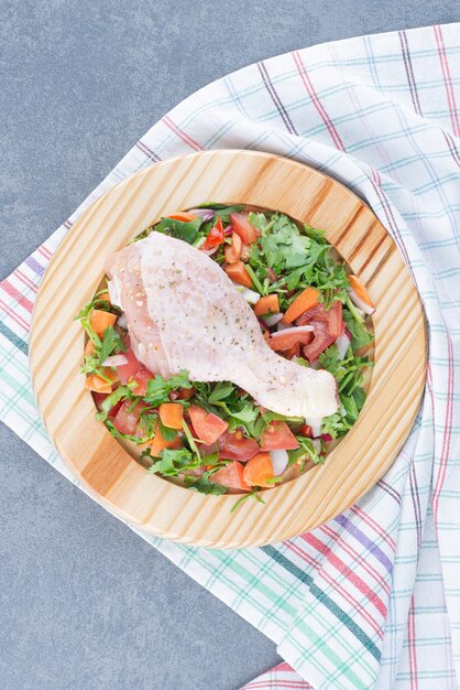 Raw chicken drumstick and sliced vegetables on wooden plate.