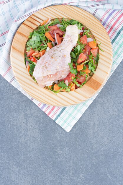 Raw chicken drumstick and sliced vegetables on wooden plate.