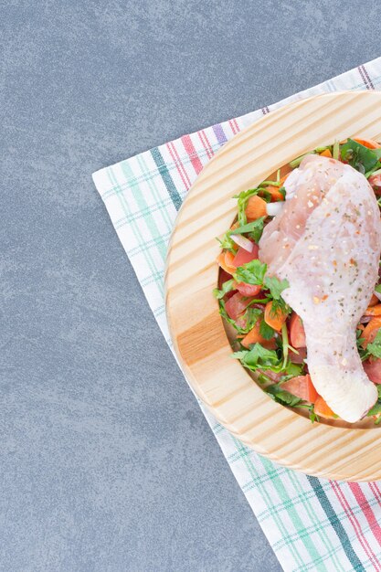 Raw chicken drumstick and sliced vegetables on wooden plate.