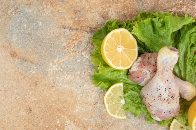 Raw chicken legs with sliced lemon and lettuce on marble surface