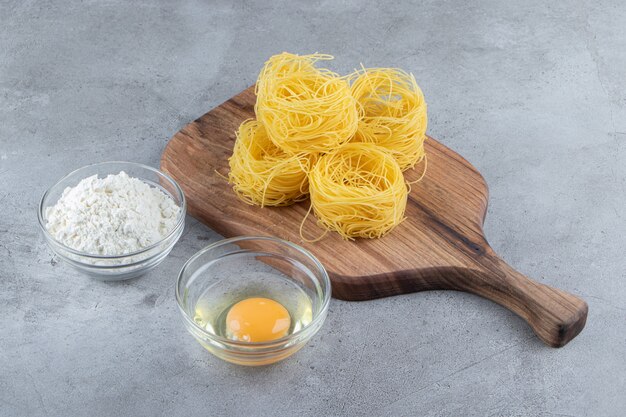 Raw dry nest pasta with raw egg and a glass bowl of flour on a stone surface .