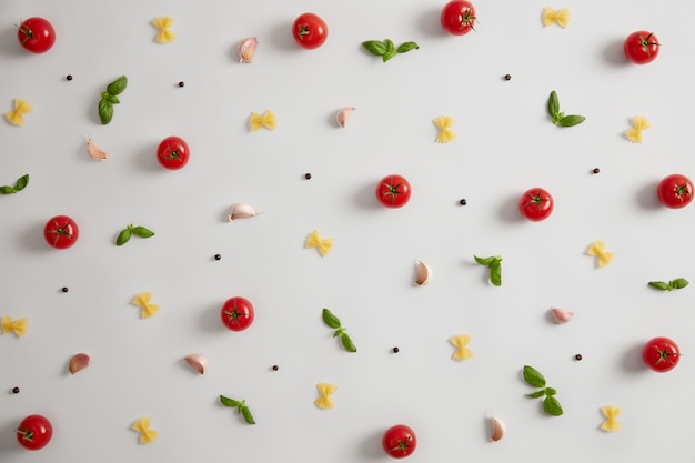 Сырые макароны в форме галстука-бабочки, красные помидоры, базилик и специи для приготовления итальянских блюд. Выборочный фокус. Макароны как источник углеводов. Традиционная кухня. Свежие сырые ингредиенты