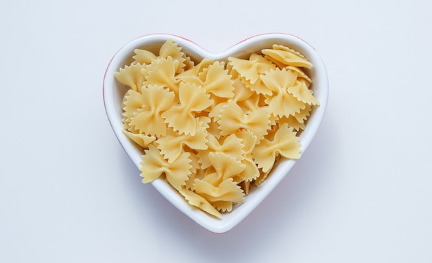 Raw farfalle pasta in a heart shaped bowl on white wall, top view.