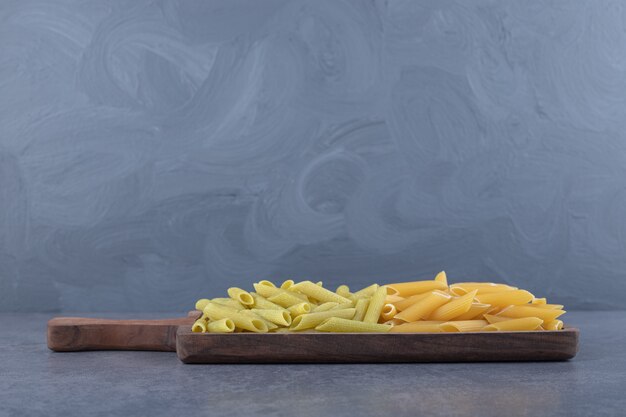 Raw green and yellow penne pasta on wooden board.