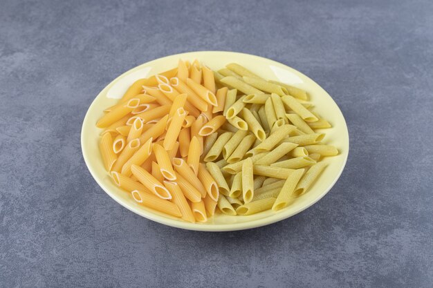 Сырые зеленые и желтые макаронные изделия пенне на желтой тарелке.