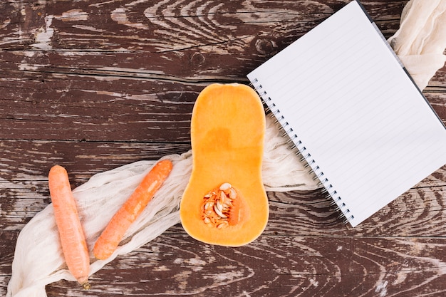 Raw halved organic butternut squash; carrots and spiral notepad on wooden table surface