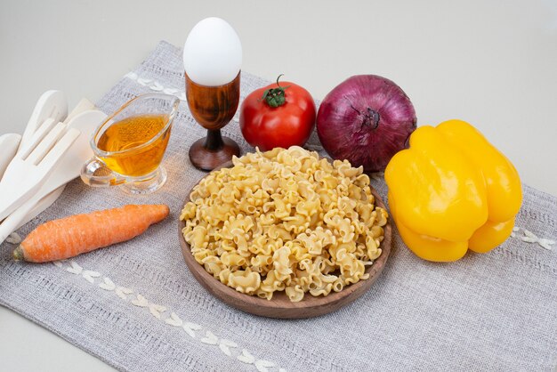 Raw macaroni with vegetables on wooden plate on tablecloth