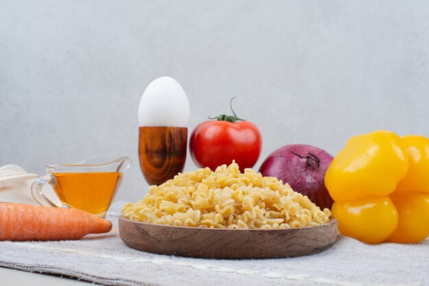 Raw macaroni with vegetables on wooden plate on tablecloth