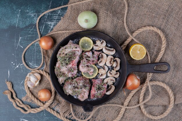Кусочки сырого мяса в черной сковороде с луком, чесноком, лимоном и грибами.