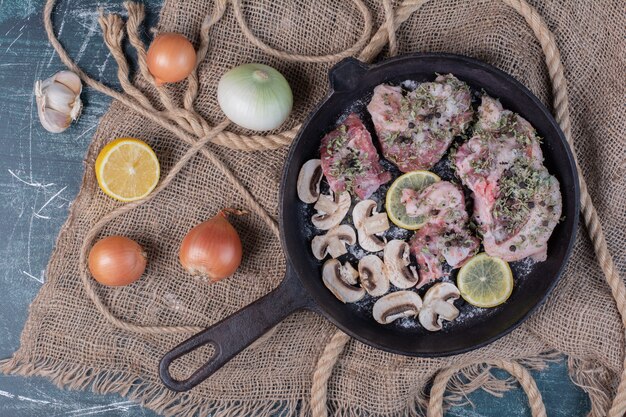 Raw meat pieces in black pan with onions, garlic, lemon and mushrooms.