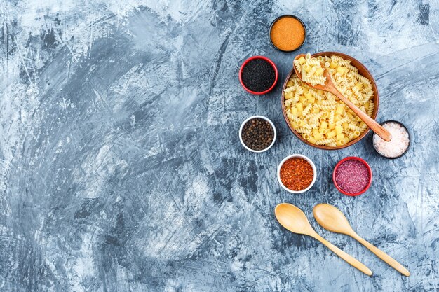 Raw pasta in a bowl with spices, wooden spoons top view on a grey plaster background