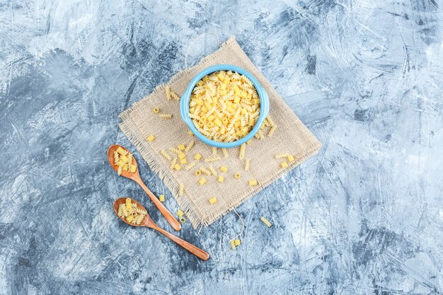 Raw pasta in bowl and wooden spoons on plaster and piece of sack background. top view.
